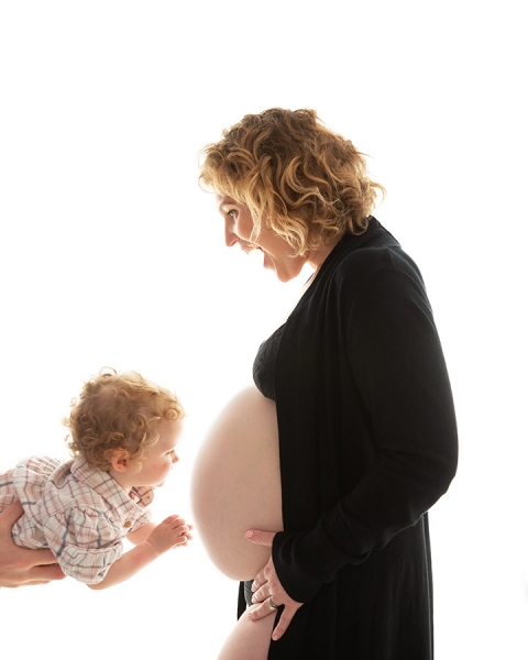 curly haired toddler in fathers hands looking at mothers bare pregnant belly